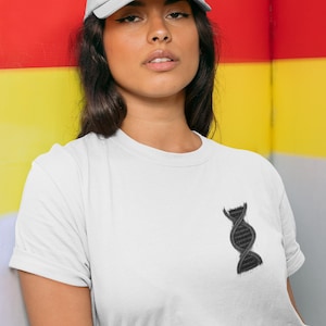 Könnte beinhalten: Eine Frau trägt ein weißes T-Shirt mit einer schwarzen DNA-Grafik und eine weiße Baseballkappe. Der Hintergrund ist eine rot-gelb-weiß gestreifte Wand.