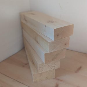 May include: A stack of five unfinished, rectangular wooden beams. The light-colored wood has visible grain and knots. The beams are stacked against a white wall and rest on a light-colored wooden surface.