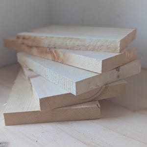 May include: A stack of five unfinished wooden boards, likely pine, with a natural wood grain. The boards are stacked on a light wood surface.
