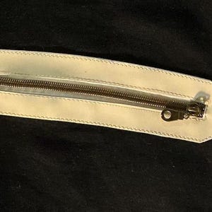 Puede incluir: Bolsillo de cuero color crema con cremallera met&aacute;lica. El bolsillo es rectangular con un borde cosido, sobre un fondo negro. La cremallera es de color met&aacute;lico oscuro.