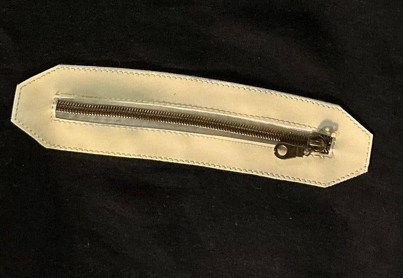 Puede incluir: Bolsillo de cuero color crema con cremallera met&aacute;lica. El bolsillo es rectangular con un borde cosido, sobre un fondo negro. La cremallera es de color met&aacute;lico oscuro.