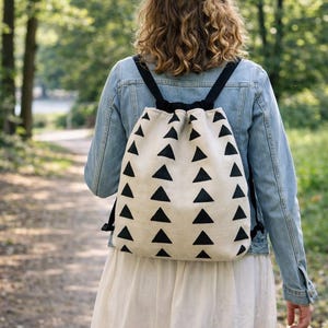 Puede incluir: Una mochila con cord&oacute;n de color crema con un patr&oacute;n repetido de tri&aacute;ngulos negros. La mochila tiene correas negras y se lleva sobre una chaqueta vaquera y un vestido blanco. El texto "ENGLISH and CZECH PDF pattern included" es visible.