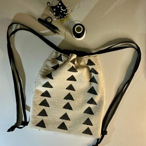 Puede incluir: Una mochila con cordón de color crema con un patrón repetido de triángulos negros. La bolsa tiene correas negras y un cierre de cordón negro. Los suministros de costura, incluyendo un alfiletero, tijeras e hilo, están colocados cerca de la bolsa.