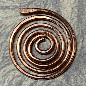 Könnte beinhalten: Ein handgefertigter Kupfer-Spiralanhänger. Der Anhänger besteht aus einer Reihe konzentrischer Kreise. Das Kupfer hat einen warmen, rötlich-braunen Farbton. Das Design ist schlicht und elegant. Der Durchmesser des Anhängers beträgt ca. 4 cm.