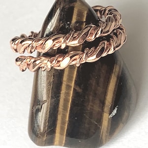 May include: A copper-coloured ring featuring a double-band design with a twisted, rope-like texture. The ring is displayed resting on a smooth, polished piece of brown and gold striped tiger's eye gemstone with a vertical orientation.