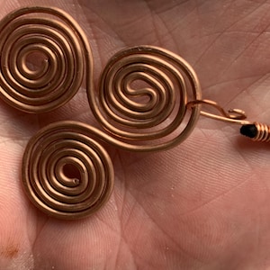 May include: Three copper spiral pendants, each with a circular design. One pendant is attached to a copper hook and black cord. The copper has a warm, reddish-brown color. The pendants are handmade and likely for jewelry.