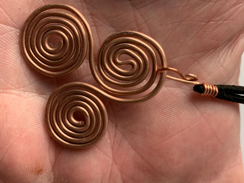 May include: Three copper spiral pendants, each with a circular design. One pendant is attached to a copper hook and black cord. The copper has a warm, reddish-brown color. The pendants are handmade and likely for jewelry.