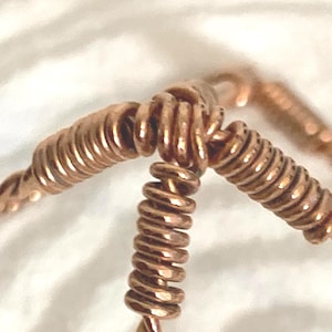 Puede incluir: Un componente de joyer&iacute;a de alambre de cobre que presenta tres segmentos distintos de alambre enrollado apretadamente que convergen en un punto central. El material de cobre met&aacute;lico muestra un tono marr&oacute;n rojizo c&aacute;lido y una textura pulida.
