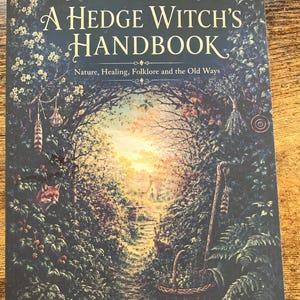 May include: A book titled The Foliate Way A Hedge Witch's Handbook Nature, Healing, Folklore and the Old Ways. The cover features a dark, lush forest path leading to a cottage, with a fox, a rabbit, a woven basket, and a wooden staff.