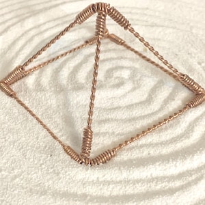 Puede incluir: Una escultura en forma de pir&aacute;mide construida con alambre de cobre trenzado. La estructura presenta cuatro esquinas en la base y un v&eacute;rtice &uacute;nico, con cada borde formado por dos hebras de alambre trenzadas. Las uniones est&aacute;n reforzadas con alambre enrollado.