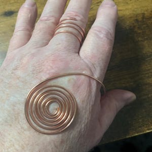 Op de afbeelding: Koperdraad ringenset. Eén ring heeft een spiraalontwerp, de andere is een meerlaagse band. De ringen worden op een hand getoond, waarbij hun ingewikkelde, handgemaakte details worden getoond. Het koper heeft een warme, roodbruine tint.