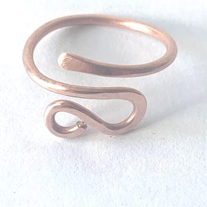 May include: A copper-colored metal ring featuring an open, bypass-style design. The band is formed from a single piece of wire that curves into a rounded, hook-like shape at one end, creating a continuous, smooth, and polished metallic surface.
