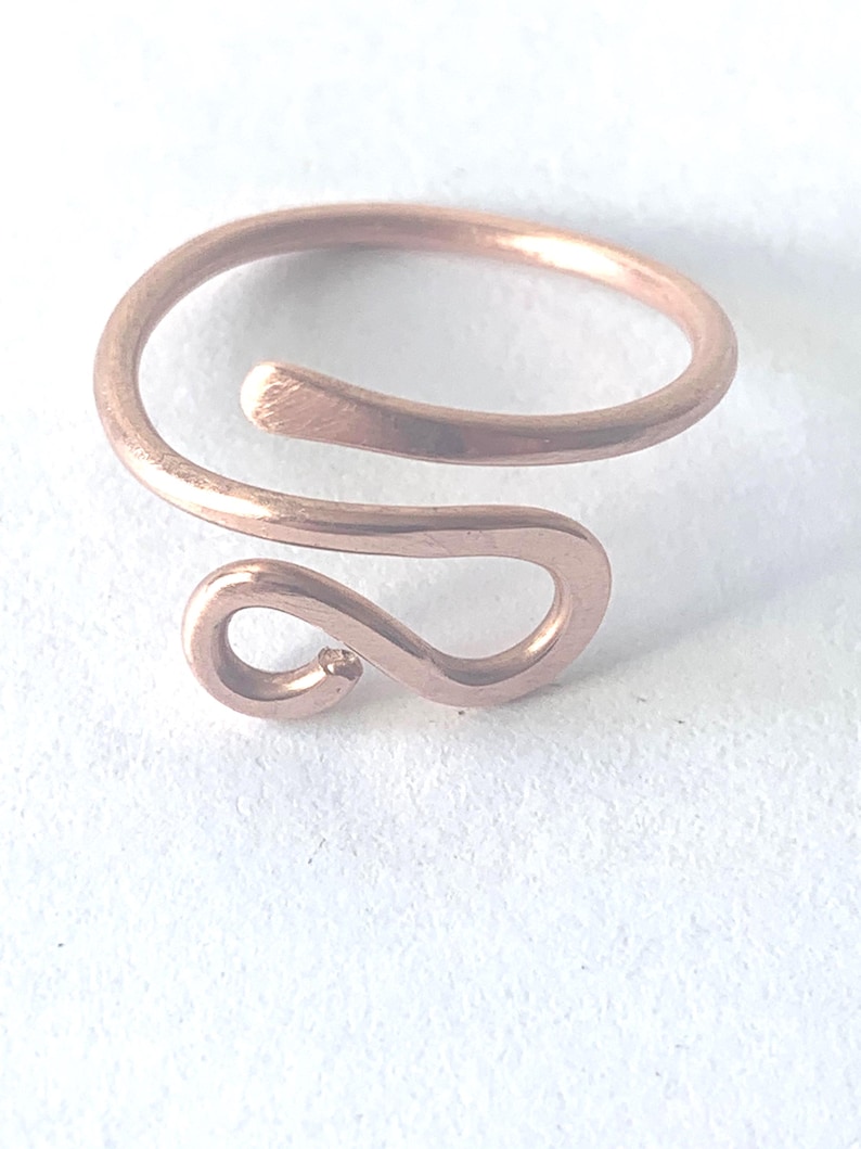 May include: A copper-colored metal ring featuring an open, bypass-style design. The band is formed from a single piece of wire that curves into a rounded, hook-like shape at one end, creating a continuous, smooth, and polished metallic surface.