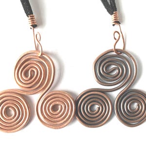 May include: Two wire pendants featuring a triple spiral design. One pendant is bright copper, and the other is a darker, oxidised copper. Each pendant is attached to a black cord with a copper wire wrap at the top, forming a hook for suspension.
