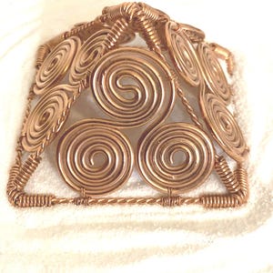 May include: A pyramid-shaped sculpture crafted from copper wire, featuring intricate spiral patterns on each face. The structure is built with twisted wire edges and coiled wire accents, creating a geometric design with a metallic, reddish-brown finish.