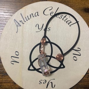 May include: A wooden divination board with the text "Arluna Celestial Yes No Yes No". A clear crystal pendulum is attached to a black ring and copper wire accents. The board has a Celtic knot design.