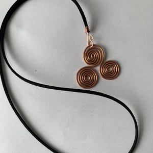May include: A copper wire pendant necklace featuring a design of three spiral circles. The pendant is attached to a black cord. The necklace is displayed on a white background.