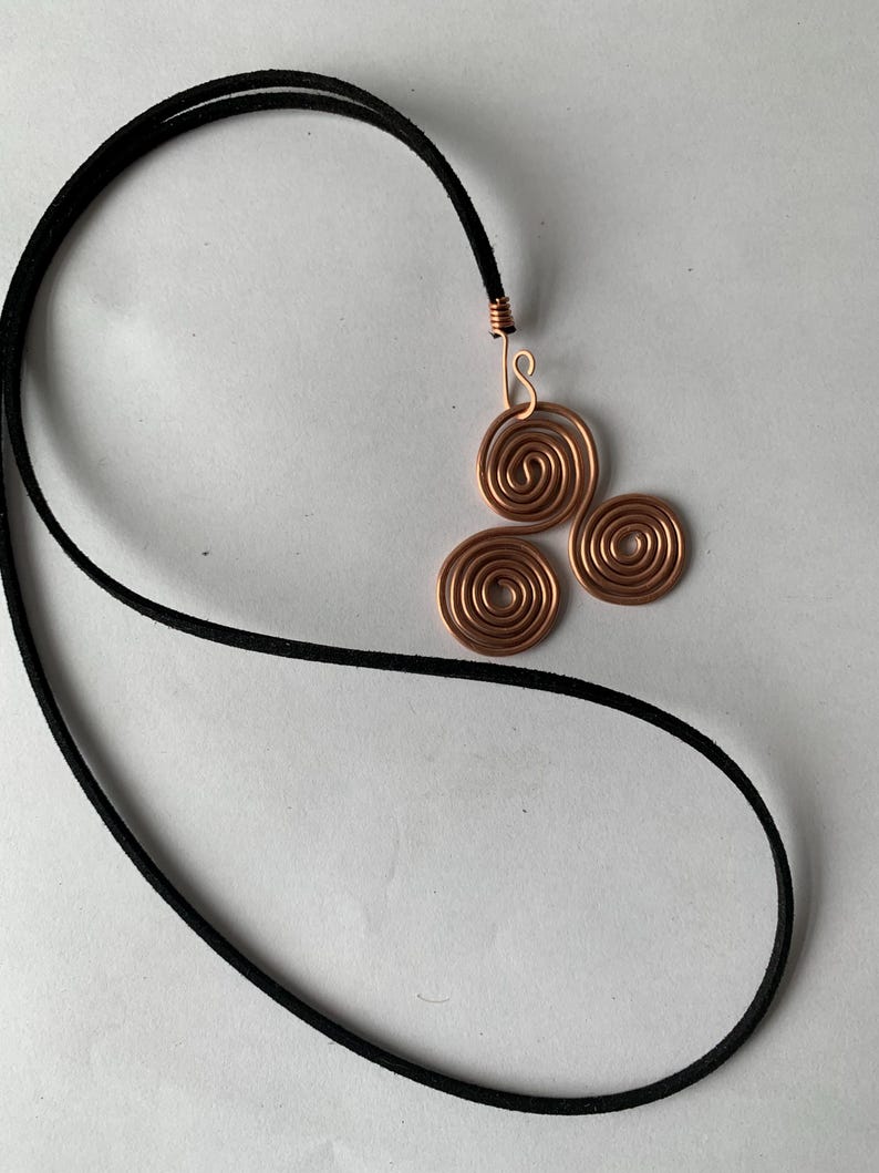 May include: A copper wire pendant necklace featuring a design of three spiral circles. The pendant is attached to a black cord. The necklace is displayed on a white background.