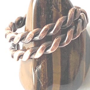 May include: A copper ring featuring a twisted, rope-like design with a dark, oxidised finish. The ring is displayed around a smooth, polished tiger's eye stone that exhibits vertical bands of golden-brown and dark brown colours.