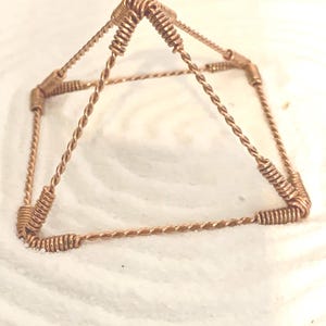 Puede incluir: Una escultura con forma de pir&aacute;mide hecha de alambre de color cobre. La estructura presenta cuatro caras triangulares que se unen en un v&eacute;rtice central, con cada borde formado por segmentos de alambre trenzado y bobinas apretadas en las esquinas.