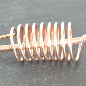 May include: A copper-coloured metal hair accessory featuring a central straight wire wrapped with a larger, wider spiral coil. The design consists of a continuous metallic structure with a polished finish, showcasing a distinct coiled spring-like shape.