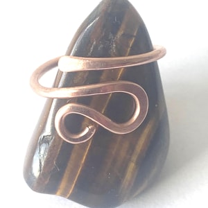 May include: A copper wire ring featuring a bypass design with a curved, open-ended loop, resting on a polished, dark brown and gold-striped tiger's eye gemstone that has a smooth, teardrop-shaped surface with natural mineral inclusions.