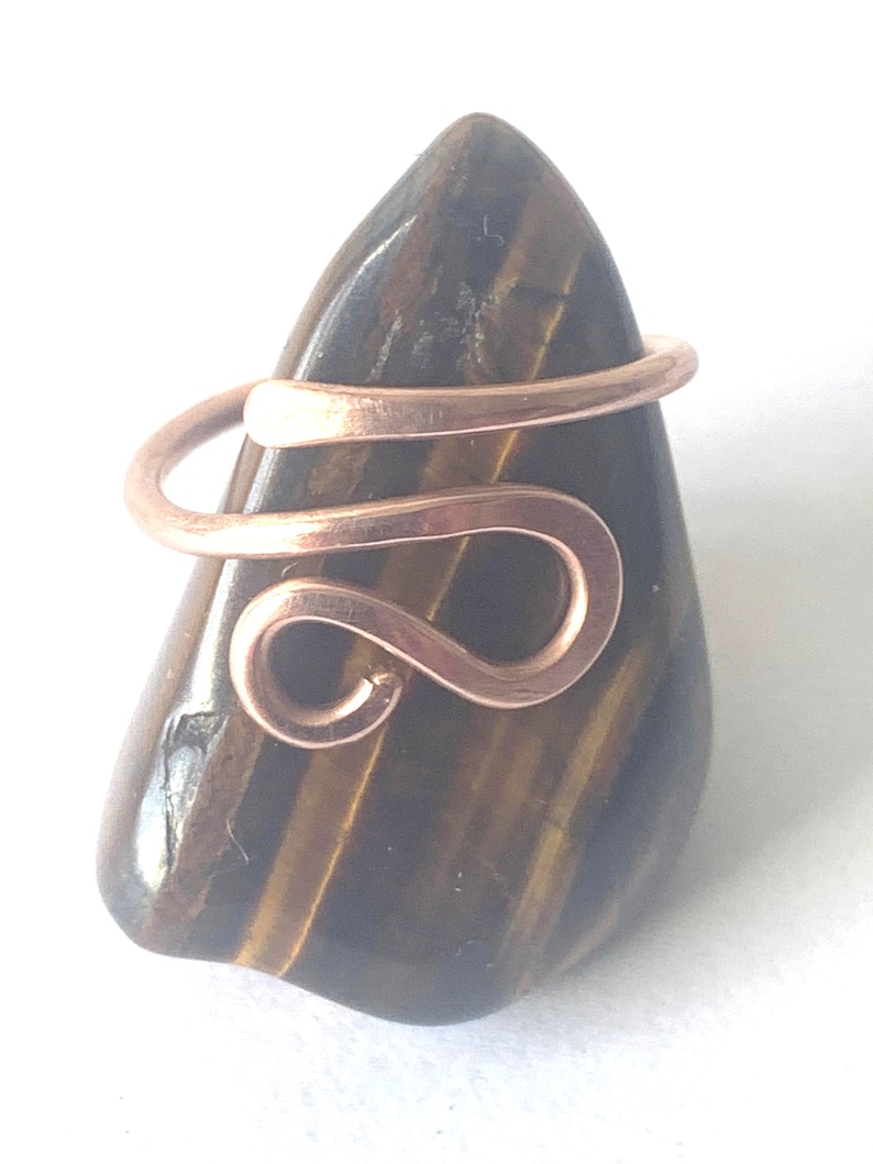 May include: A copper wire ring featuring a bypass design with a curved, open-ended loop, resting on a polished, dark brown and gold-striped tiger's eye gemstone that has a smooth, teardrop-shaped surface with natural mineral inclusions.