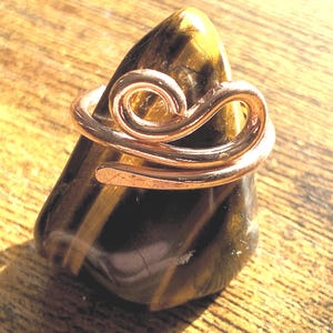 May include: A copper wire ring featuring a swirling, open-ended design wrapped around a polished, triangular-shaped tiger's eye gemstone. The stone displays characteristic golden-brown and dark brown bands with a smooth, reflective surface.