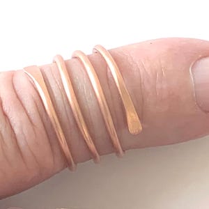 May include: A rose gold-coloured metal ring featuring a continuous spiral design that wraps around the finger. The band is composed of a single wire that coils three times, with each end tapering to a smooth, flattened finish on the surface.