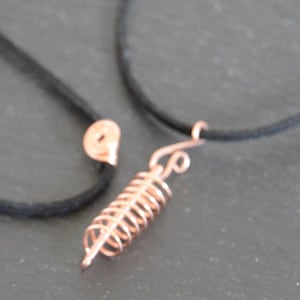 May include: A necklace featuring a black cord and a copper-coloured wire pendant shaped like a coiled spring. The pendant is attached to the cord with a small wire loop, and a separate small copper-coloured spiral component is visible nearby.
