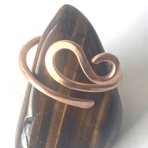 May include: A copper wire ring featuring a swirling, open-ended design wrapped around a polished, teardrop-shaped tiger's eye gemstone. The metal has a warm, metallic finish, while the stone displays characteristic brown and gold bands.