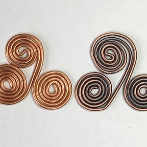 May include: Two copper wire ornaments, each with three connected spiral shapes. One is a solid copper colour, and the other has a darker, almost black, patina. The ornaments are displayed against a white background.