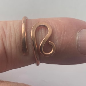 May include: A copper-coloured ring with an open design. The ring features a swirling, abstract shape on the top and a simple band that wraps around the finger. The ring is made of a shiny, metallic material.