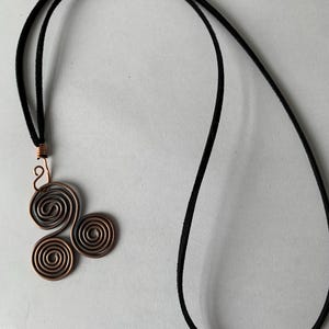 May include: A copper-colored pendant necklace featuring a cluster of three spiral designs. The pendant is attached to a black cord necklace. The spirals are made of copper wire, and the necklace cord is a dark, velvety material.