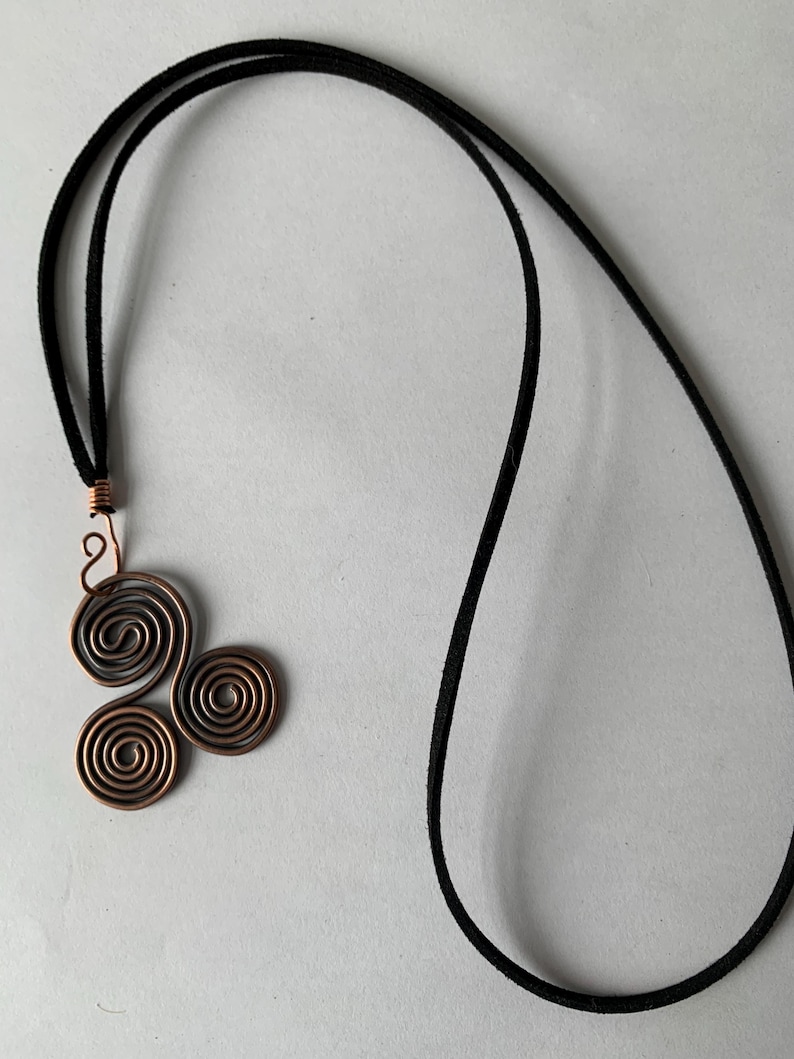 May include: A copper-colored pendant necklace featuring a cluster of three spiral designs. The pendant is attached to a black cord necklace. The spirals are made of copper wire, and the necklace cord is a dark, velvety material.