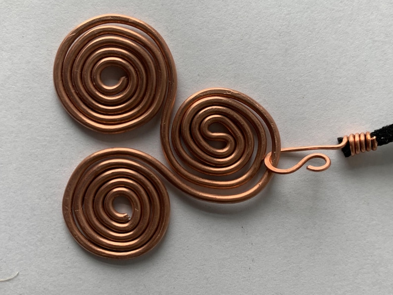 May include: A copper-colored pendant featuring three spiral designs connected by wire. The pendant is attached to a black cord with a copper clasp. The spirals are made of coiled wire.