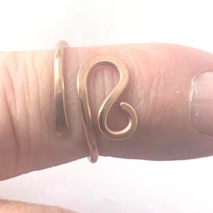 May include: A rose gold-toned metal ring featuring an open, bypass-style design. The band is formed from a single piece of wire that curves into a stylized, rounded loop shape at the front, with smooth, polished surfaces and a warm metallic finish.