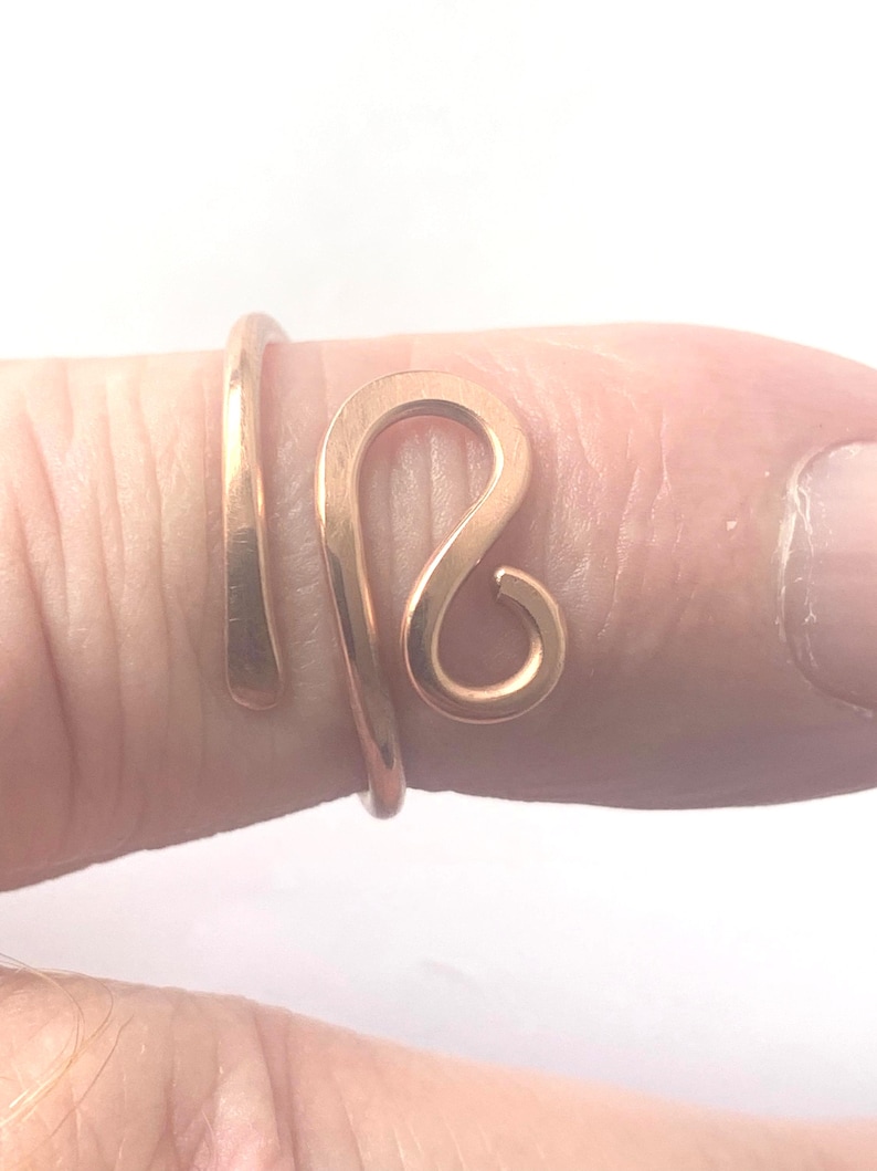 May include: A rose gold-toned metal ring featuring an open, bypass-style design. The band is formed from a single piece of wire that curves into a stylized, rounded loop shape at the front, with smooth, polished surfaces and a warm metallic finish.