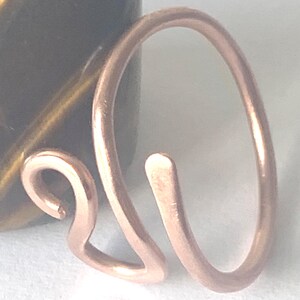 May include: A copper-colored metal ring featuring an open, bypass-style design. The band is formed from a single piece of wire that curves into a rounded shape on one side and terminates in a small, decorative loop on the other side.