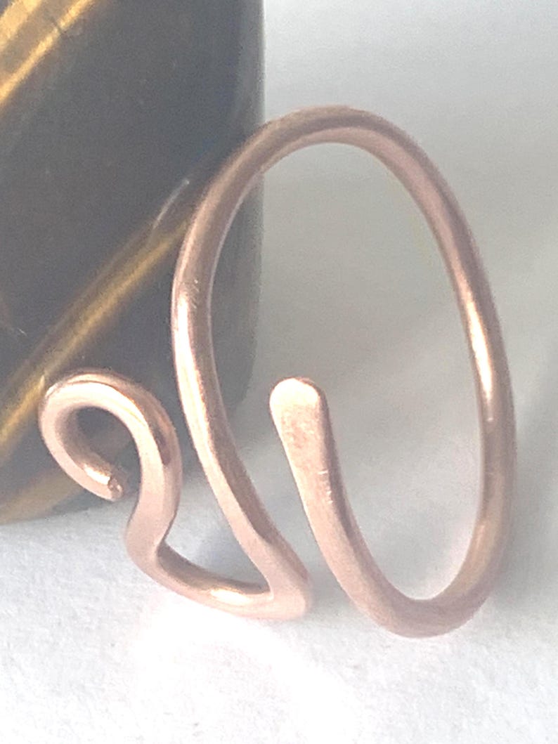 May include: A copper-colored metal ring featuring an open, bypass-style design. The band is formed from a single piece of wire that curves into a rounded shape on one side and terminates in a small, decorative loop on the other side.