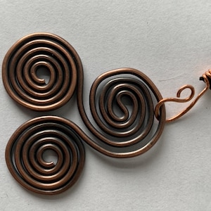 May include: A copper-colored pendant necklace featuring three spiral designs. The pendant is connected to a black cord with a copper wire clasp. The spirals are made of copper wire.