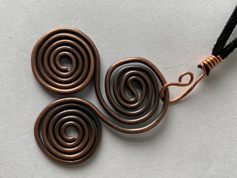 May include: A copper-colored pendant necklace featuring three spiral designs. The pendant is connected to a black cord with a copper wire clasp. The spirals are made of copper wire.