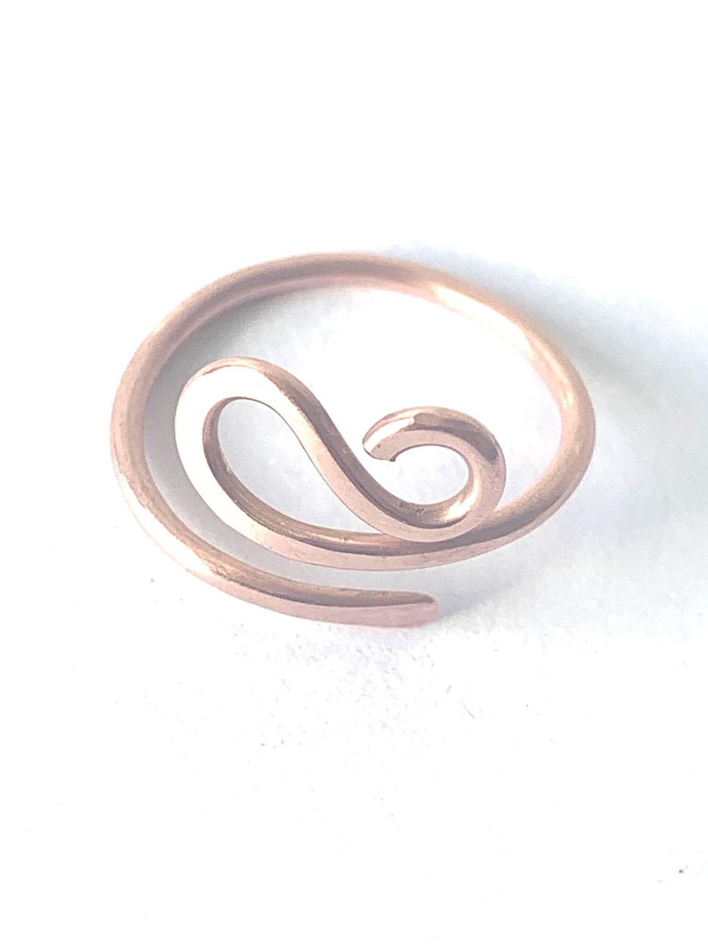 May include: A rose gold-toned metal ring featuring a continuous, open-ended design that curves into a central swirl. The band has a smooth, polished finish and a circular shape that remains open at one end, creating a fluid, wave-like aesthetic.