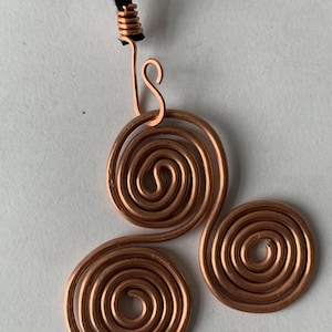 May include: A copper wire pendant necklace featuring three spiral designs. The pendant is attached to a black cord. The spirals are made of copper wire and have a shiny finish. The necklace is a handmade piece of jewelry.