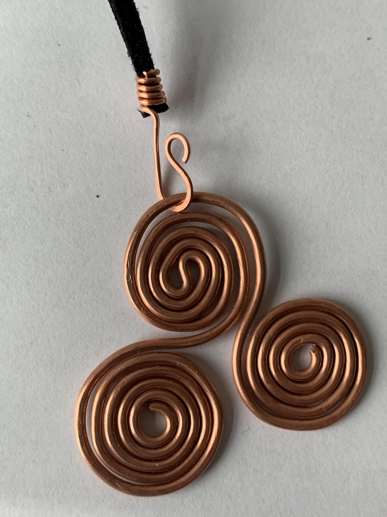 May include: A copper wire pendant necklace featuring three spiral designs. The pendant is attached to a black cord. The spirals are made of copper wire and have a shiny finish. The necklace is a handmade piece of jewelry.