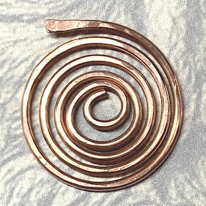 May include: A circular pendant crafted from a single length of polished copper wire, shaped into a continuous, multi-layered spiral pattern. The metal surface exhibits a warm, metallic reddish-brown hue with subtle hammered textures along the wire.