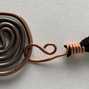 May include: Close-up of a copper pendant with a spiral design. The pendant is connected to a black cord. The copper has a natural, aged appearance. The spiral is a prominent feature.