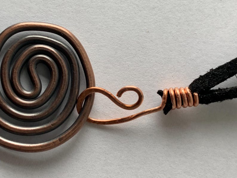 May include: Close-up of a copper pendant with a spiral design. The pendant is connected to a black cord. The copper has a natural, aged appearance. The spiral is a prominent feature.