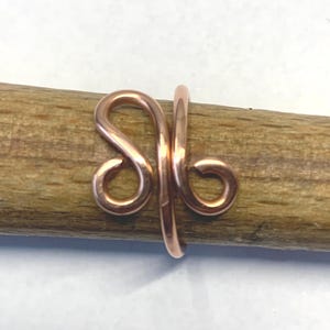 May include: A copper-coloured metal ring featuring a continuous wire design that forms two symmetrical, swirling loops on the front. The band is crafted from a single piece of polished wire, creating an open, fluid shape with rounded ends.