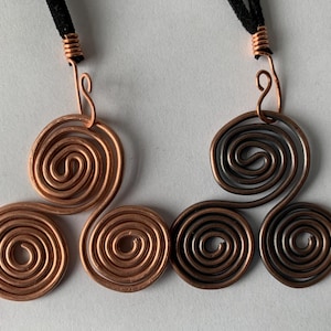May include: Two copper wire spiral pendants with black cords. One pendant is a bright copper color, and the other is a darker, oxidized copper. Each pendant features three connected spirals, with a loop at the top for the cord.
